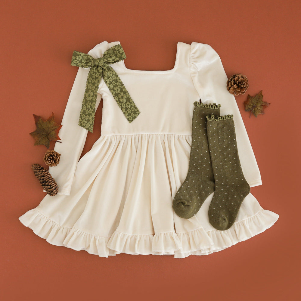 An ivory long-sleeve dress with a ruffled hem and a green patterned bow on one sleeve, styled flat-lay with matching green patterned socks, pinecones, and autumn leaves on a terracotta background.