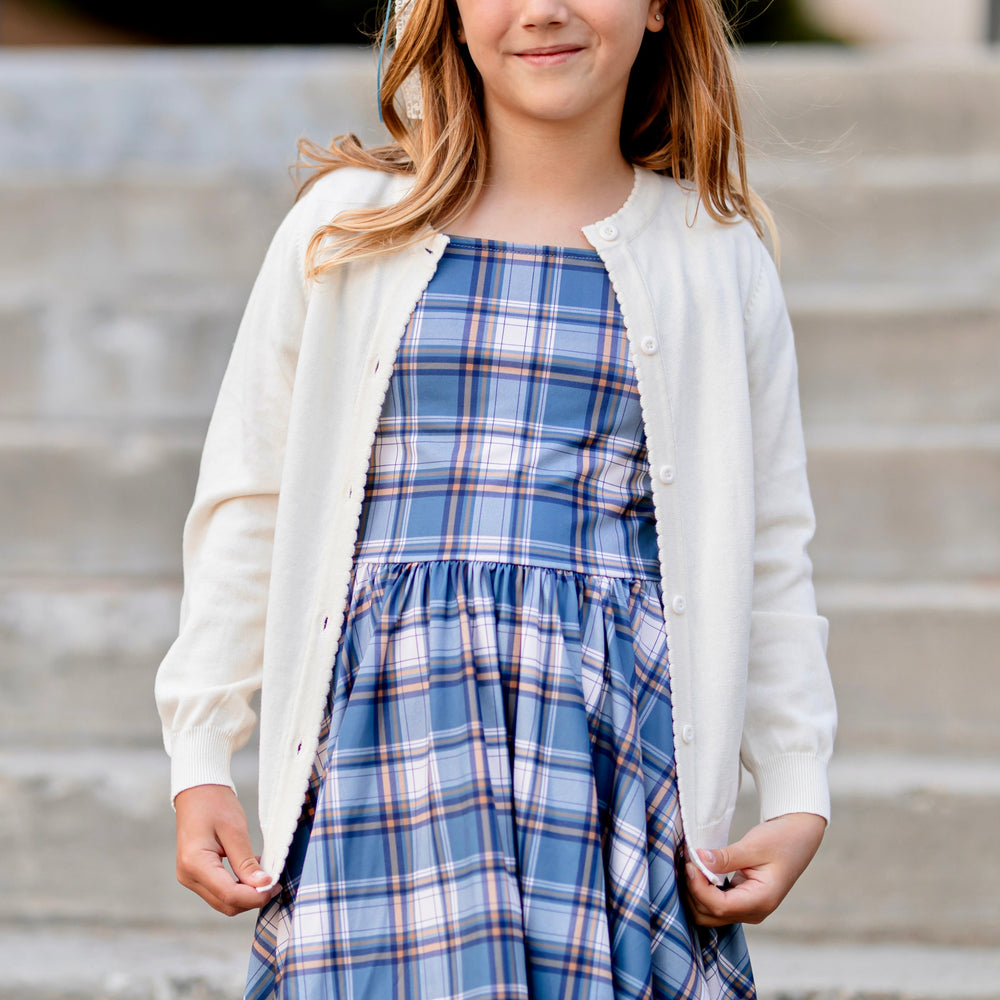 girl in blue plaid twirl dress layered with light cream button front cotton knit cardigan with scalloped trim