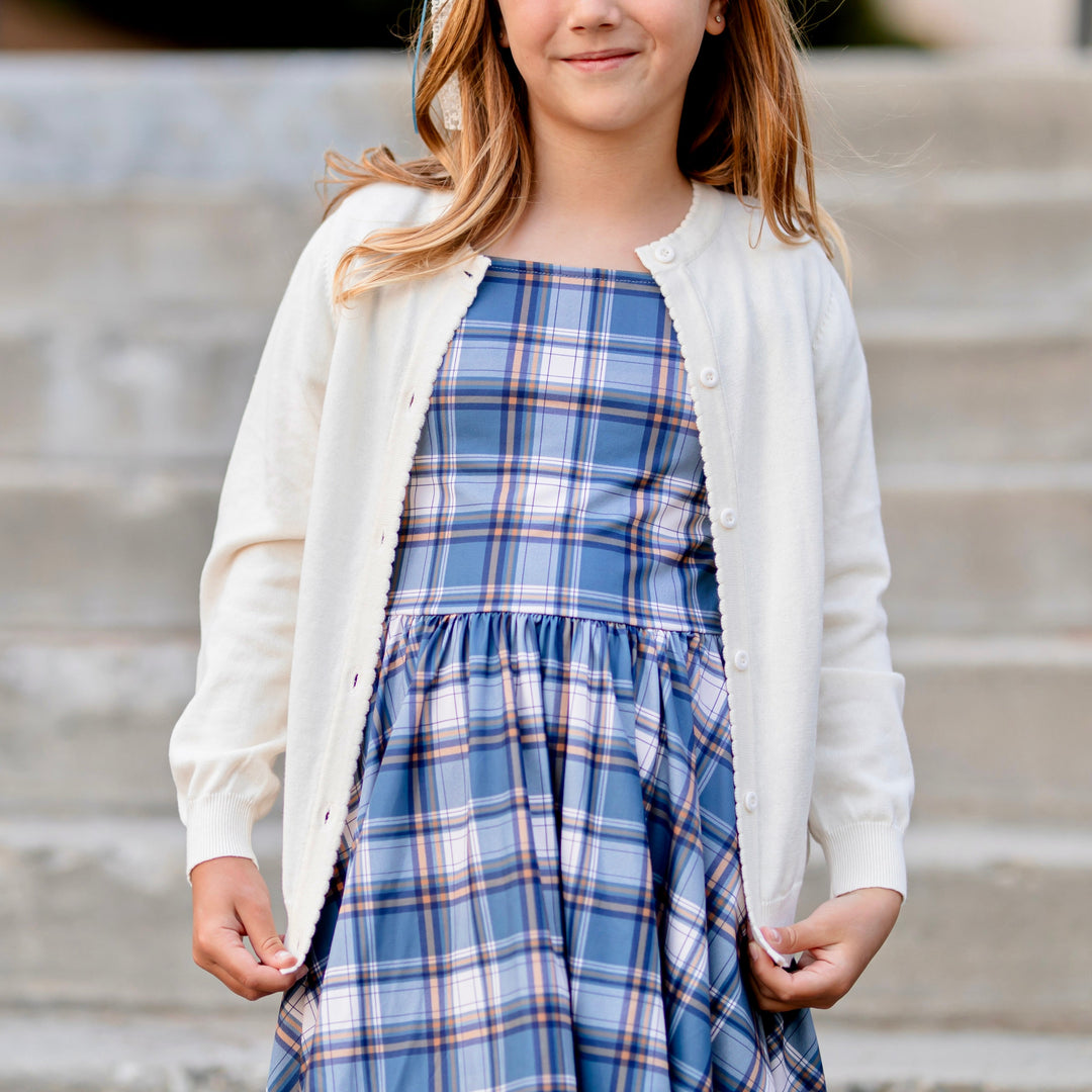 girl in blue plaid twirl dress layered with light cream button front cotton knit cardigan with scalloped trim