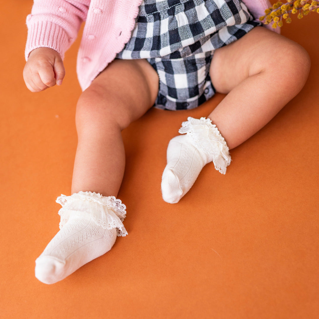 baby girl wearing ivory lace trim bobby socks with black and white buffalo plaid romper and pink knit sweater