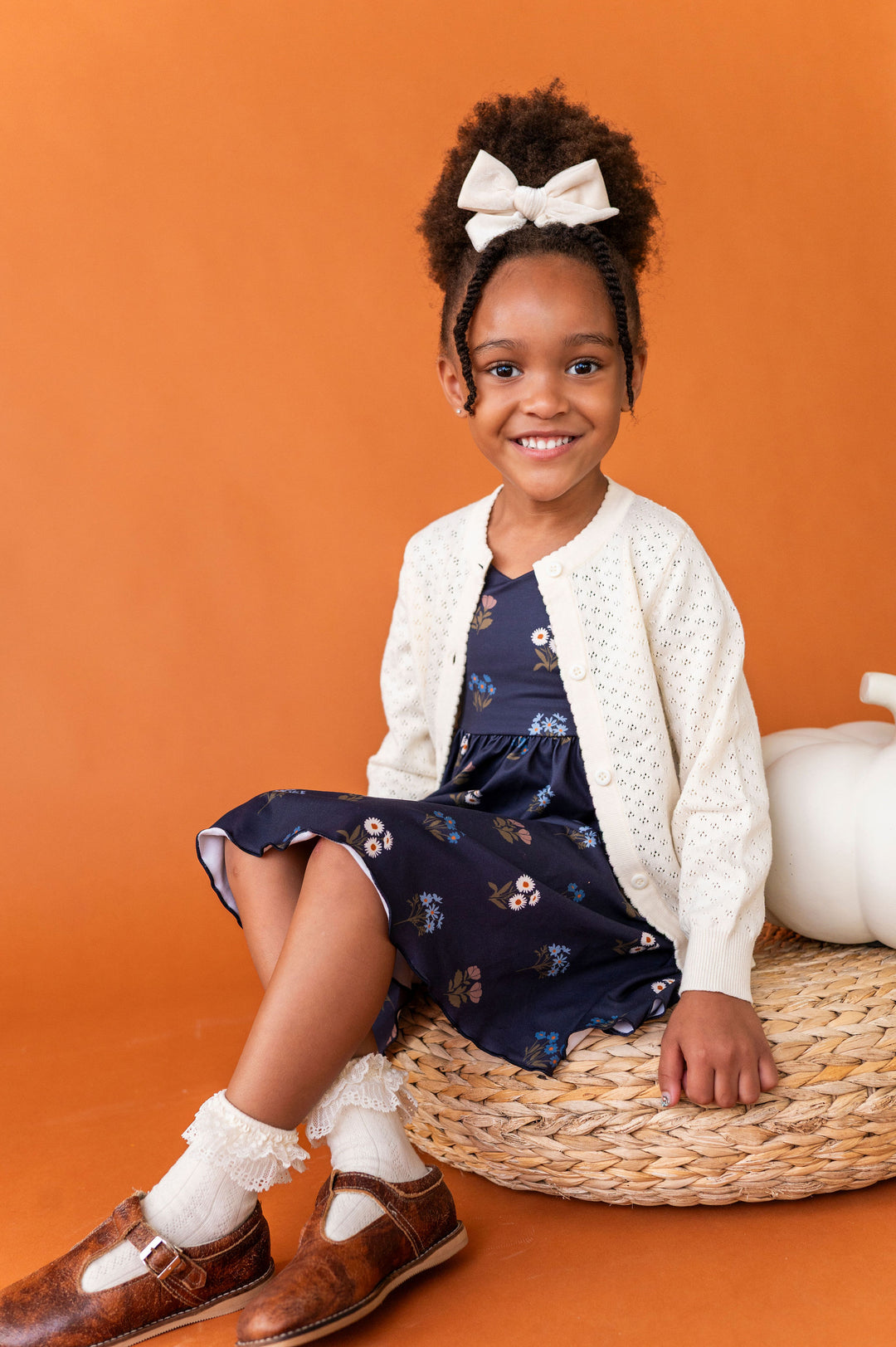 A young girl with her hair in a high bun and a white bow sits on a woven round basket against a solid orange background. She is wearing a navy blue floral dress, an ivory textured cardigan, ivory ruffled socks, and brown leather shoes.