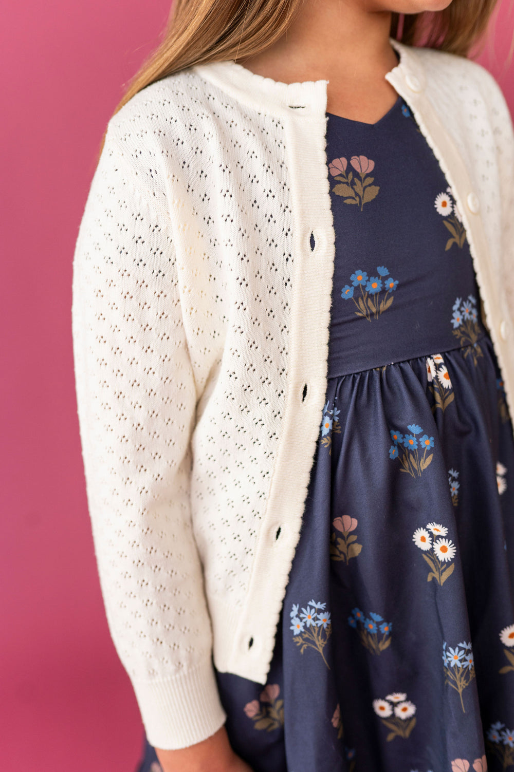 A close-up of a girl wearing an ivory open-knit cardigan over a navy blue floral dress, set against a solid pink background.
