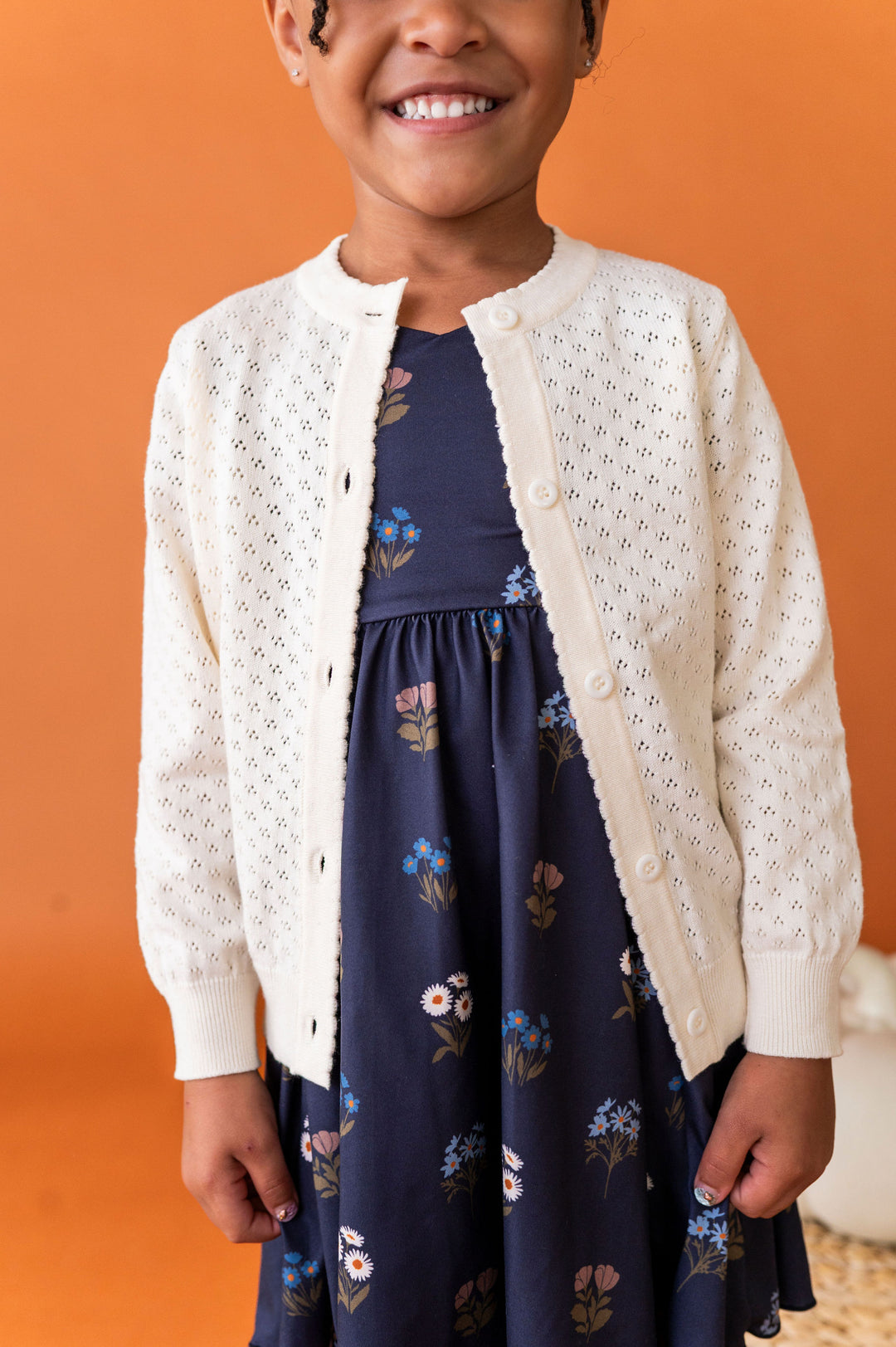 A smiling young girl wearing an ivory colored knit cardigan over a navy blue floral dress, standing against a solid orange background.