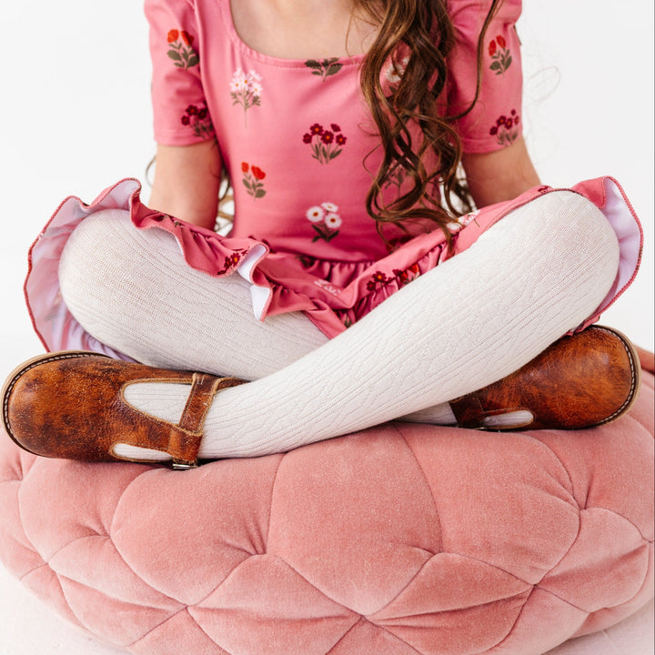 little girl sitting on pink velvet cushion wearing ivory cable knit tights paired with brown mary janes and pink floral fall twirl dress