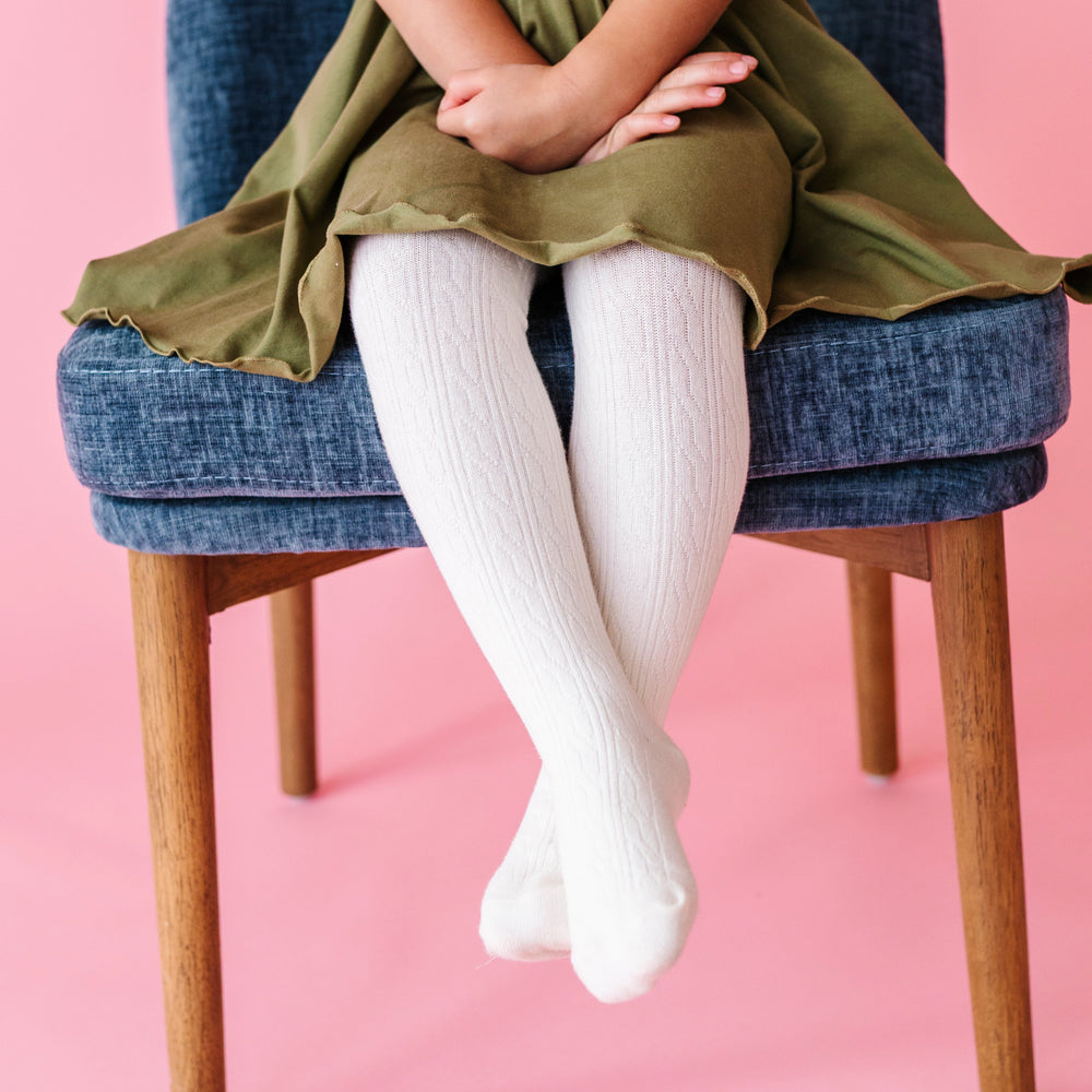 young girl sitting on chair in ivory cable knit tights paired with olive tiwlr dress