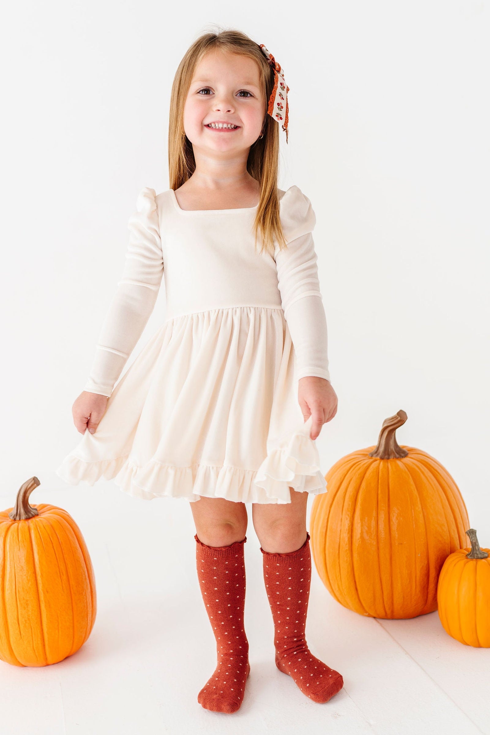 girl wearing ivory velvet dress with rust polka dot knee high socks standing with pumpkins