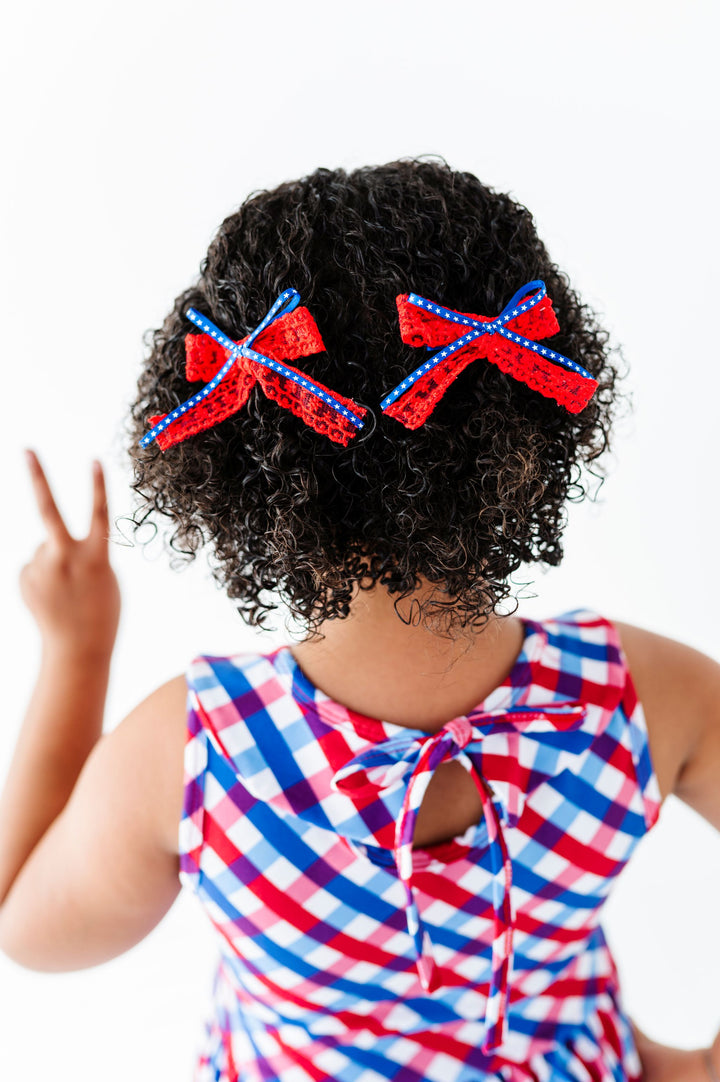 little girl wearing red, white and blue gingham 4th of July dress paired with red lace and blue star ribbon pigtail bows