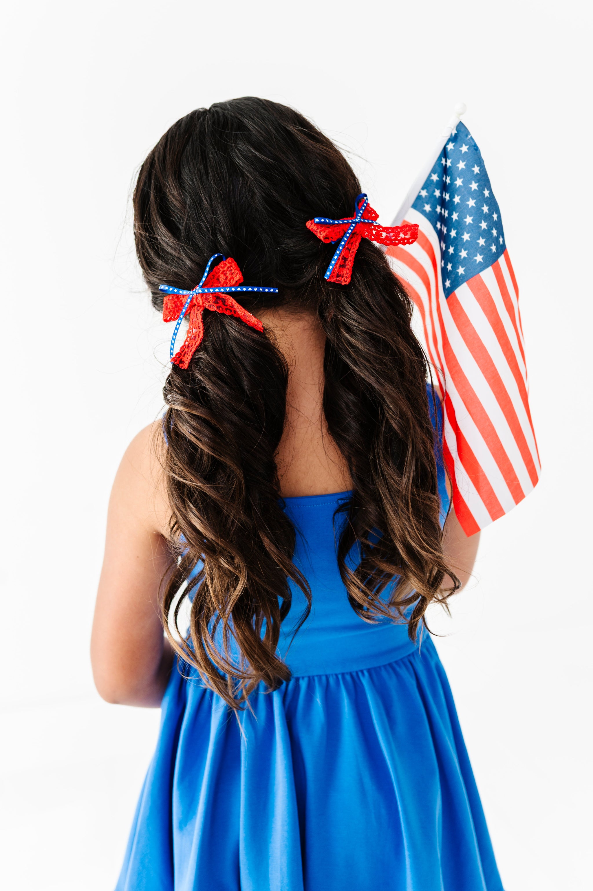 little girl holding American flag in royal blue sleeveless twirl dress and red lace and blue star ribbon pigtail bows