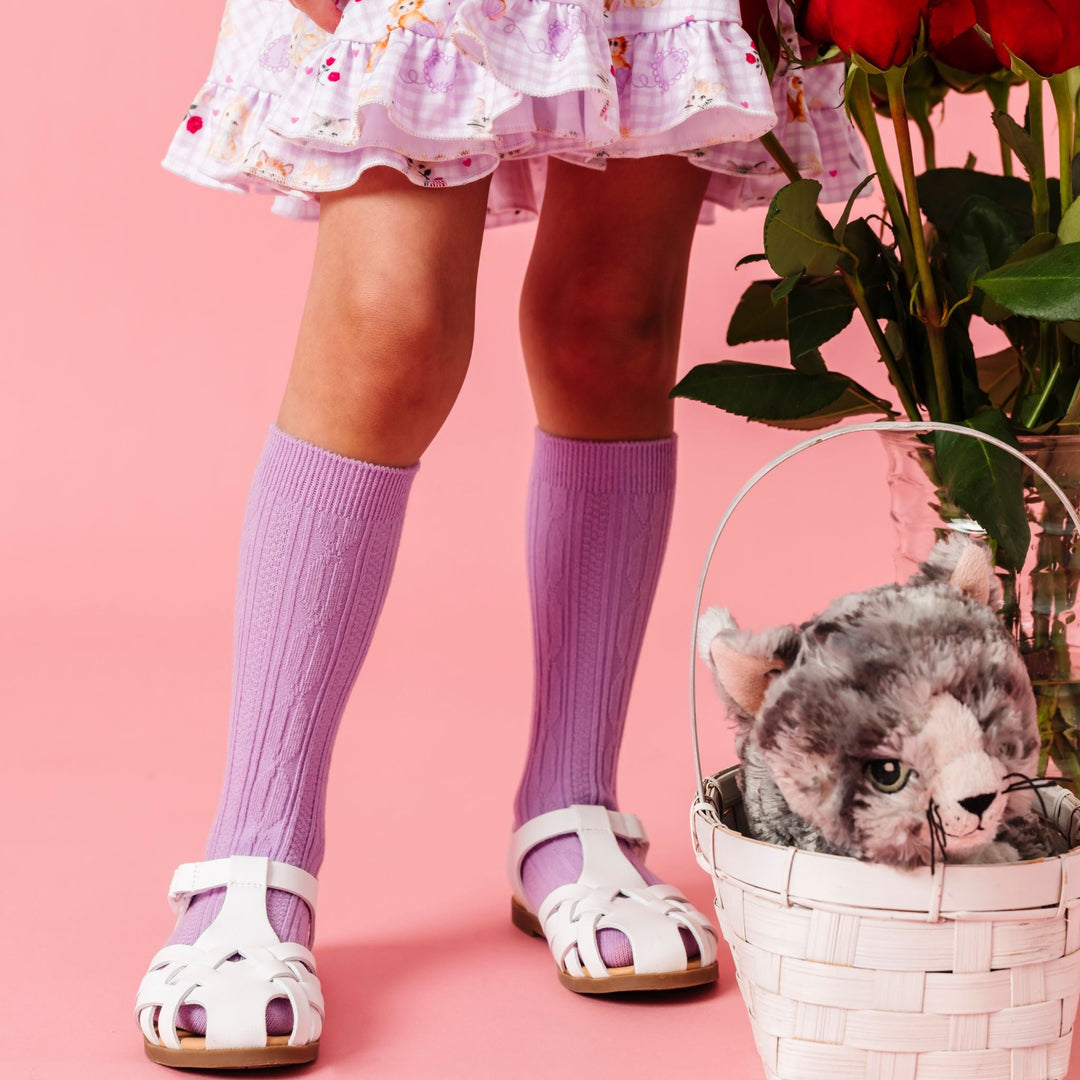 girl standing by red roses and stuffed kitten in basket wearing lavender cable knit knee socks with white sandals and matching lavender gingham kitten print twirl dress