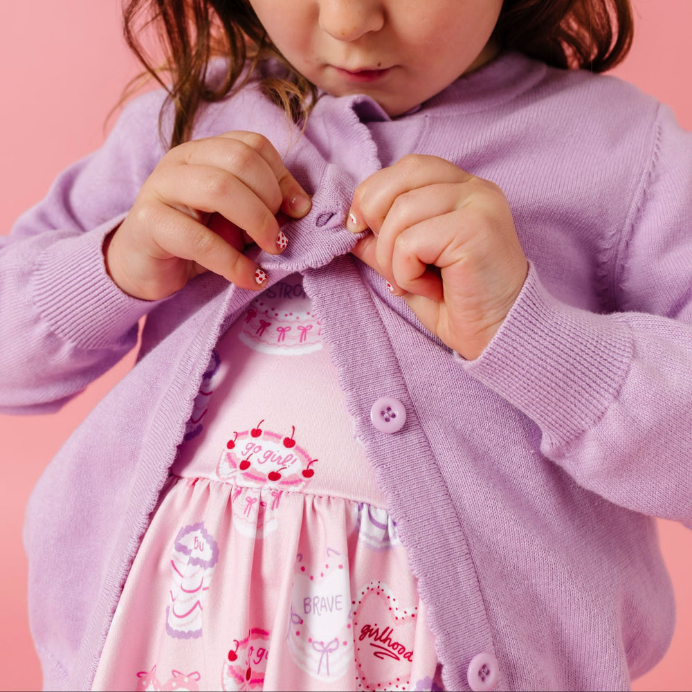 little girl buttoning light purple cotton knit cardigan over pink twirl dress with valentine's cakes and white lace hair bow