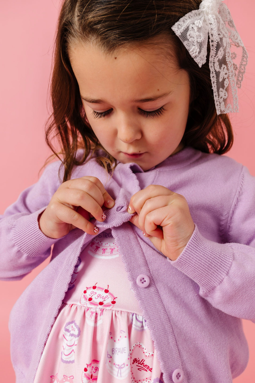 little girl buttoning light purple cotton knit cardigan over pink twirl dress with valentine's cakes and white lace hair bow
