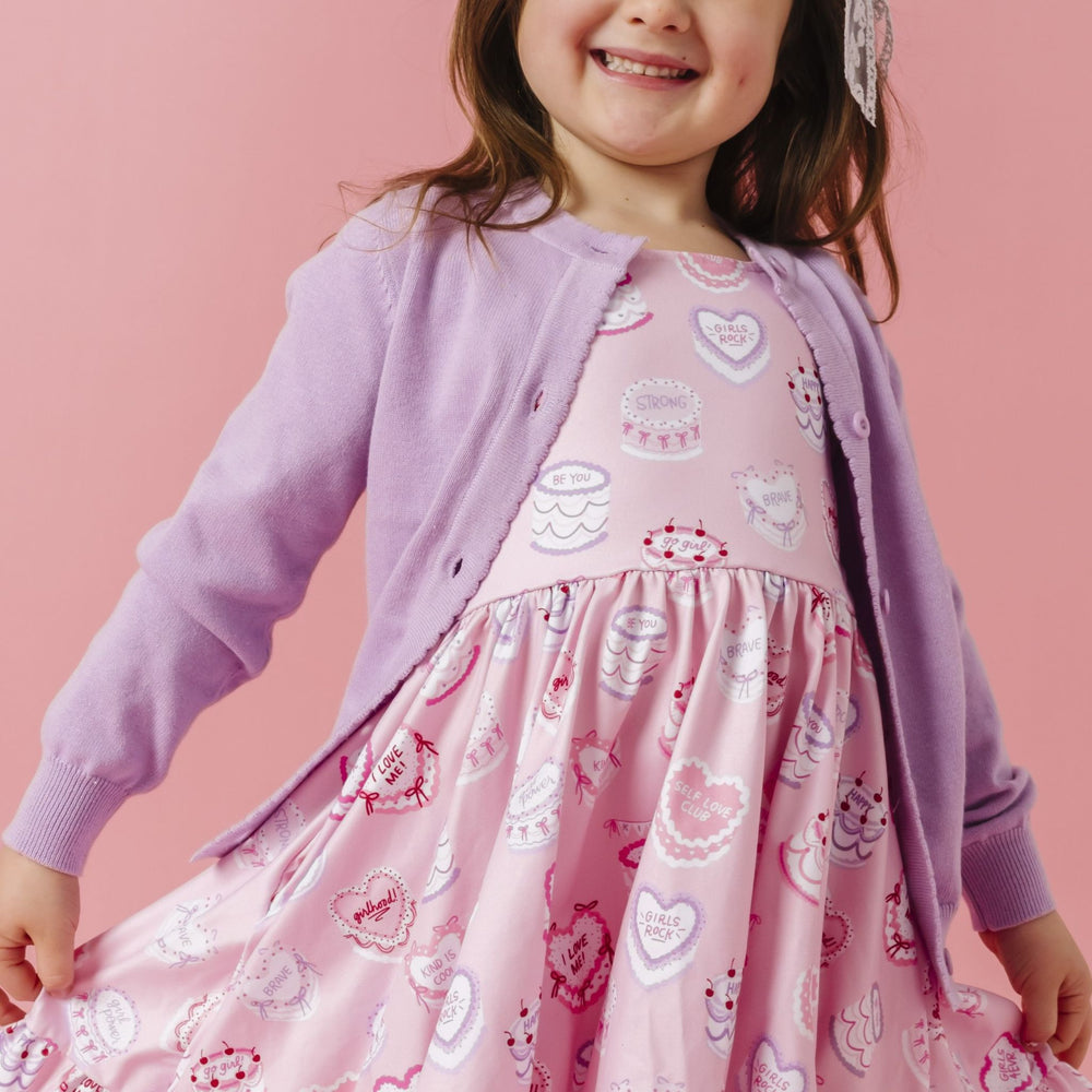 little girl in light purple button front knit cardigan with scalloped trim paired with pink twirl dress with cakes and words of affirmation