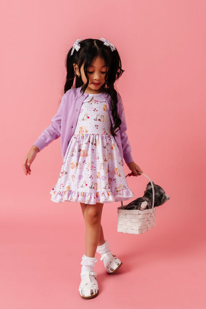 little girl holding stuffed cat in basket wearing vintage kitten print twirl dress with lavender gingham background paired with lavender cardigan, white lace bobby socks and white lace bows with lavender ribbon