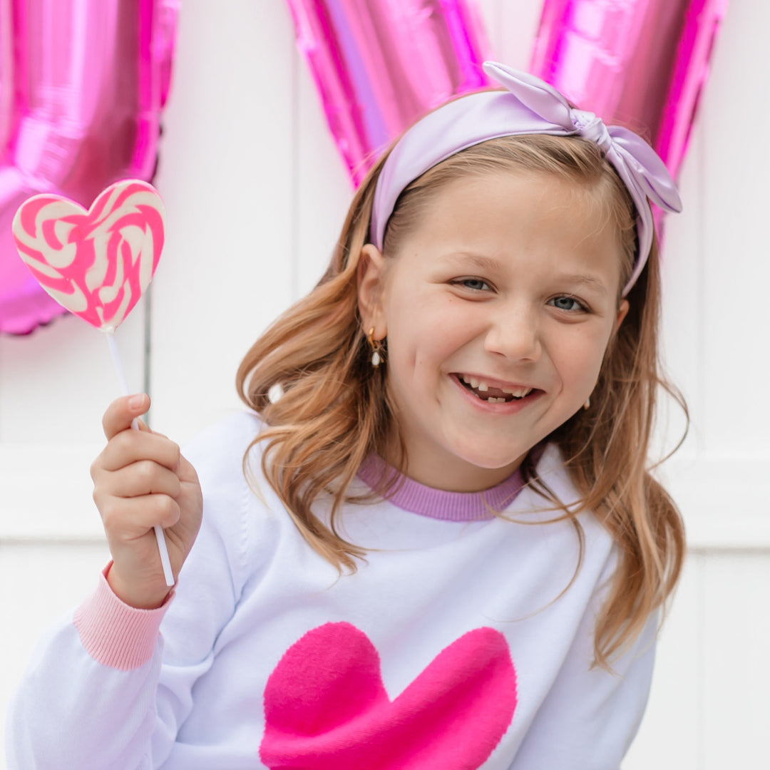 little girl holding heart sucker in lavender satin bow headband and matching Valentine's Day heart knit sweater