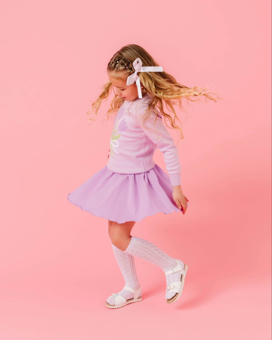 girl twirling in lavender cotton blend skort paired with matching tulip sweater, white open knit knee socks and white satin hair bow