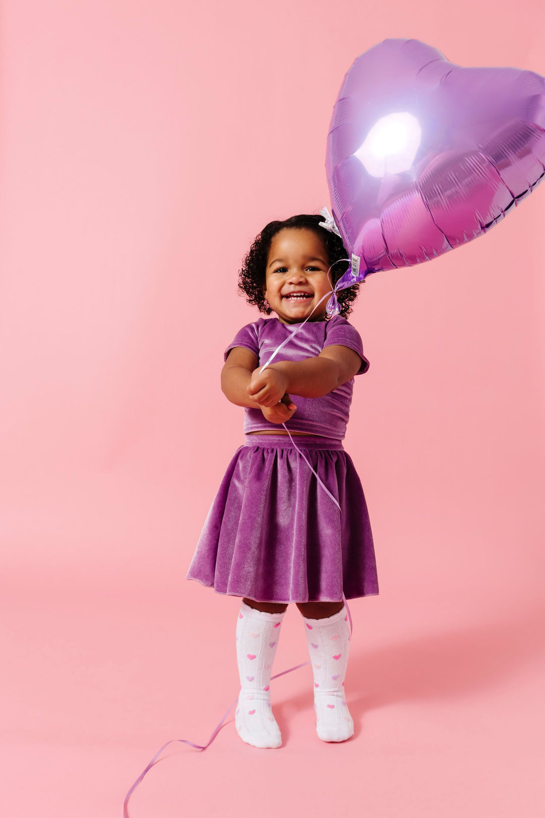 little girl holding purple heart balloon in matching lavender velvet top and skort set paired with white scalloped knee high socks with pastel hearts
