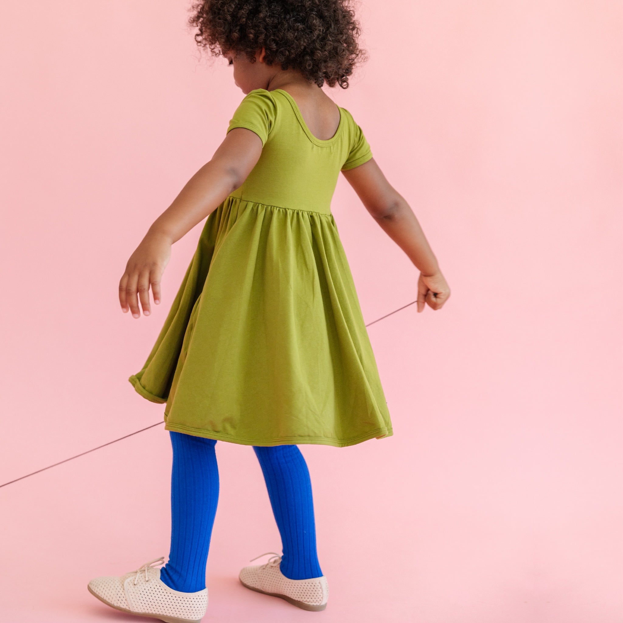 little girl wearing bright olive green cotton pocket dress and bright blue ribbed tights