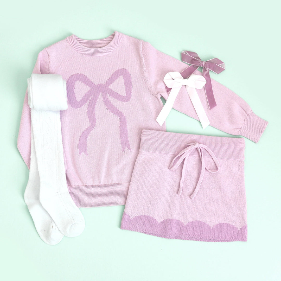 flatlay of girls spring outfit with lilac bow sweater and matching knit skirt with contrast scallop detail and velvet bows