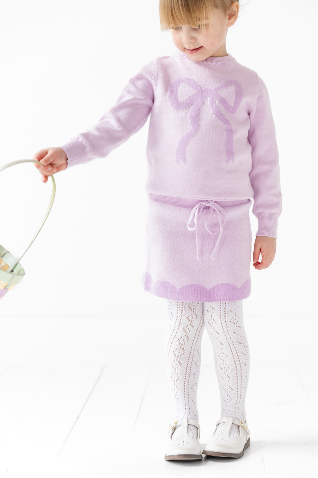 girl in light purple cotton knit pullover sweater with purple bow design with matching drawstring skirt with contrasting scallop design and wearing white openknit fancy tights