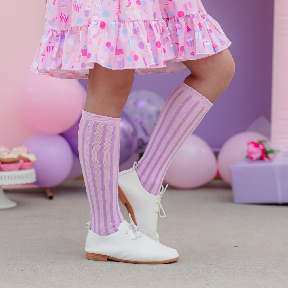 girl in lilac and lavender striped knee highs with scallop top standing in front of birthday scene and wearing matching girls birthday dress