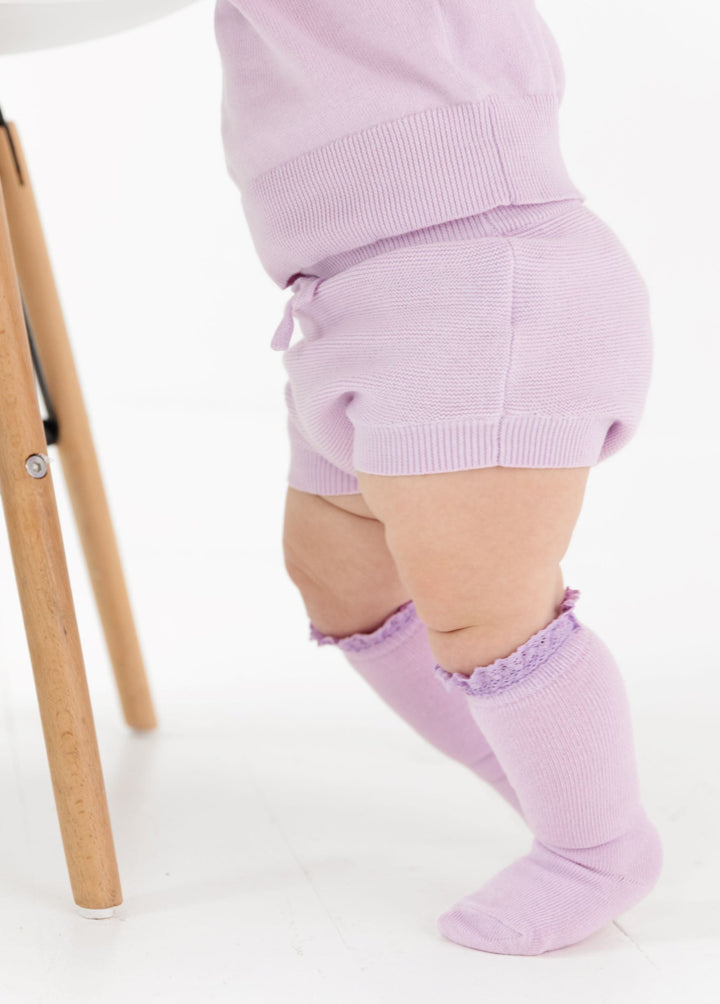 baby girl standing in lilac cotton knit bloomers with matching sweater and lace top knee highs for spring