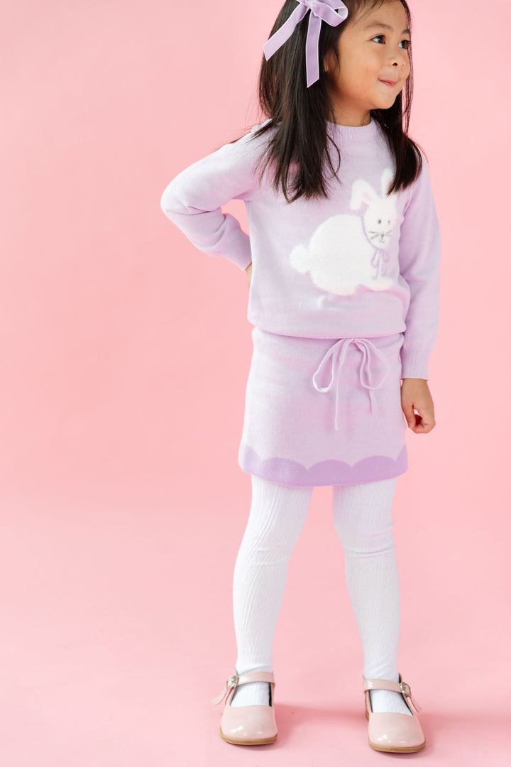 little girl in pastel purple sweater set featuring pullover knit sweater with white bunny design, sweater skirt with contrast scalloped trim, white tights and purple velvet bow