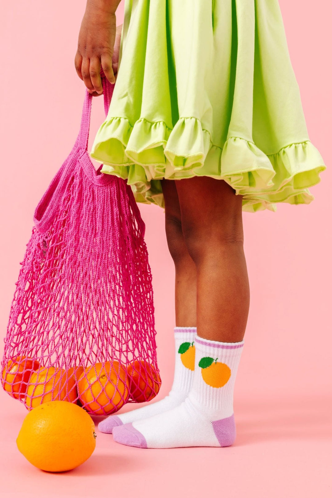 little girl holding bag of oranges in white crew socks with orange design paired with lime green summer twirl dress