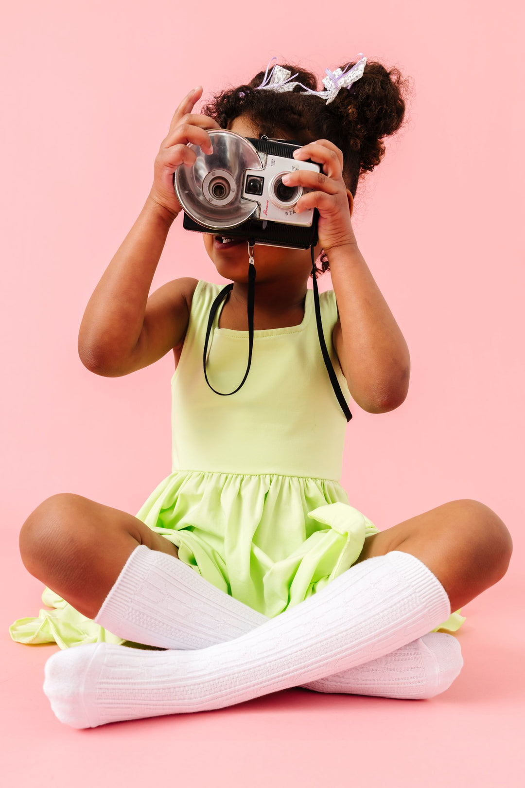 little girl with vintage camera in lime green sleeveless twirl dress paired with white cable knit knee socks