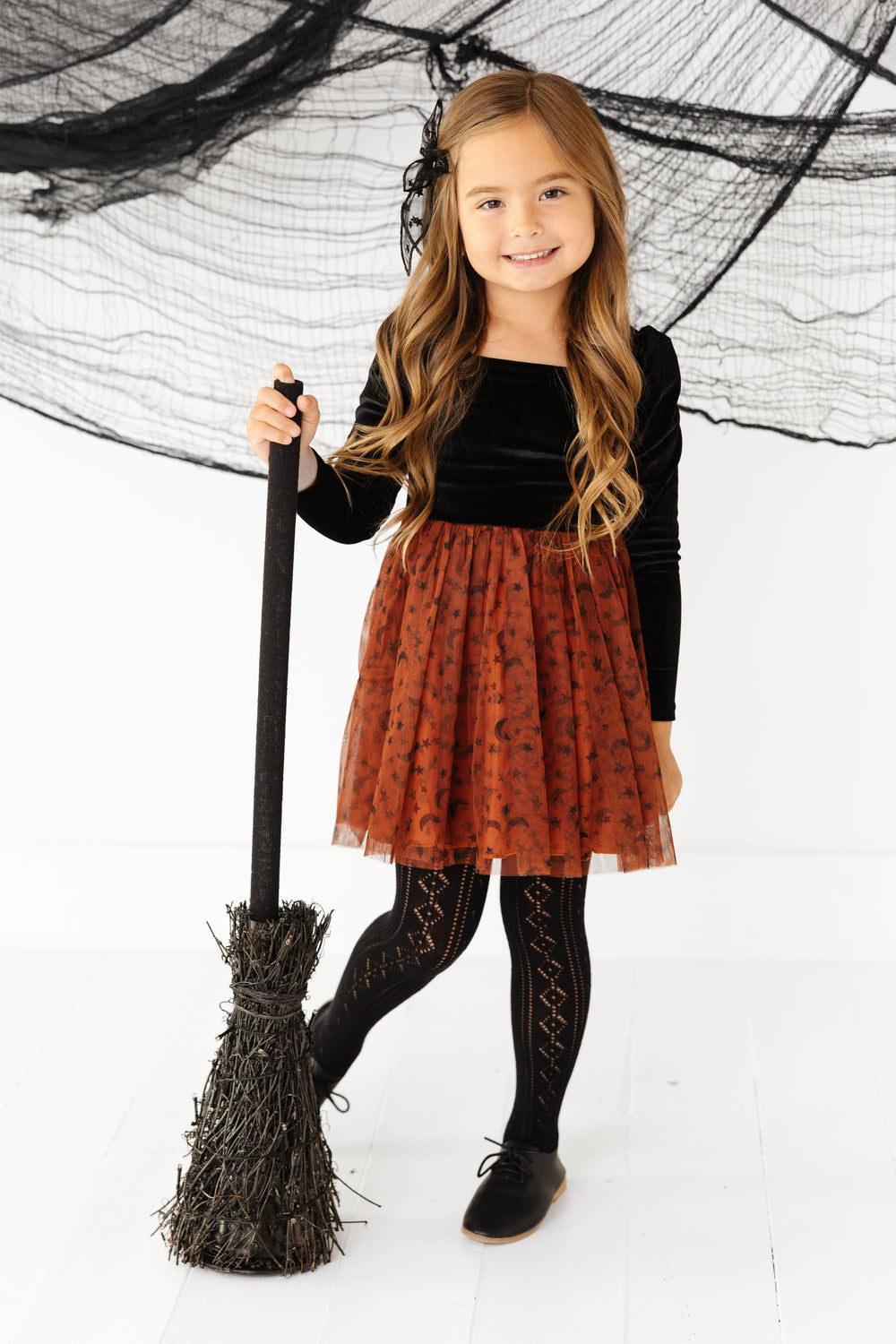 little girl holding witch broom in velvet tutu dress with black velvet long sleeve bodice and sugar almond tulle skirt with moon and star print paired with black open knit tights