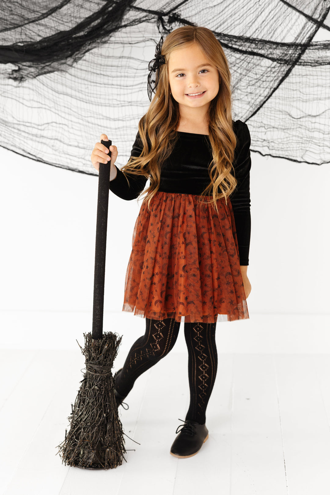 little girl holding witch broom in velvet tutu dress with black velvet long sleeve bodice and sugar almond tulle skirt with moon and star print paired with black open knit tights