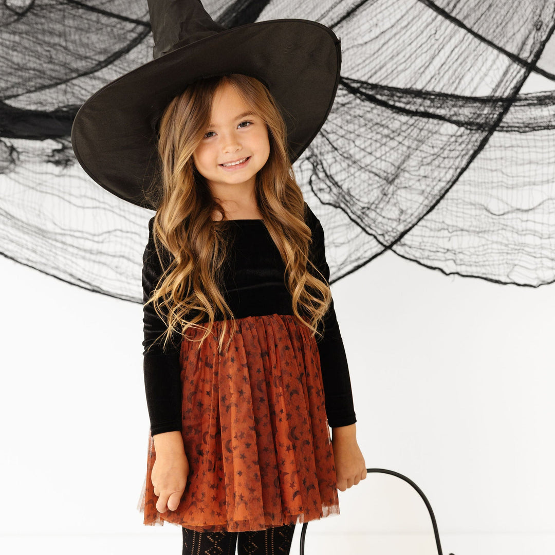 girl in Halloween witch costume dress with witch hat, tutu, and black shoes holding a cauldron.