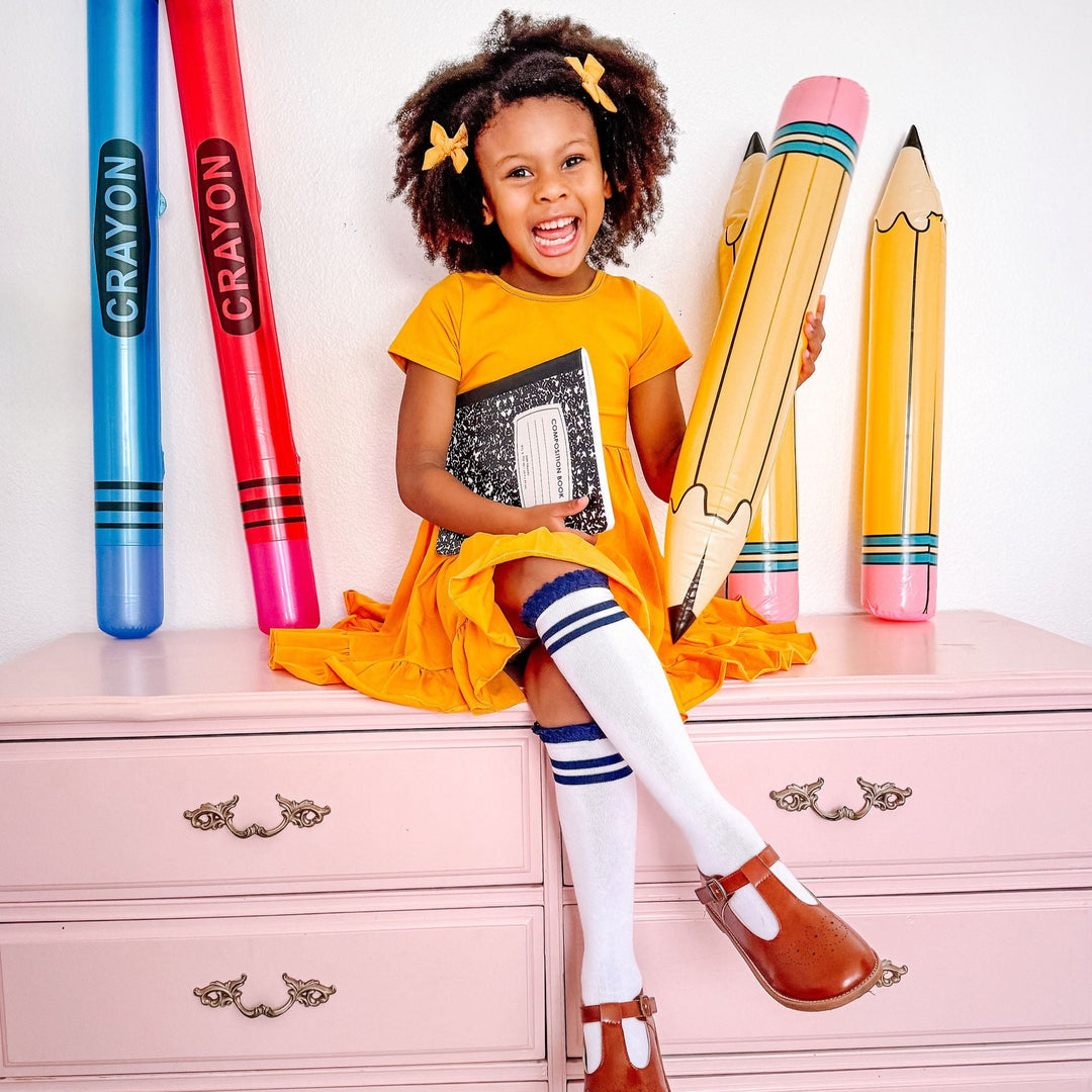 little girl sitting on pink dresser wearing marigold yellow dress with navy and white striped knee high socks