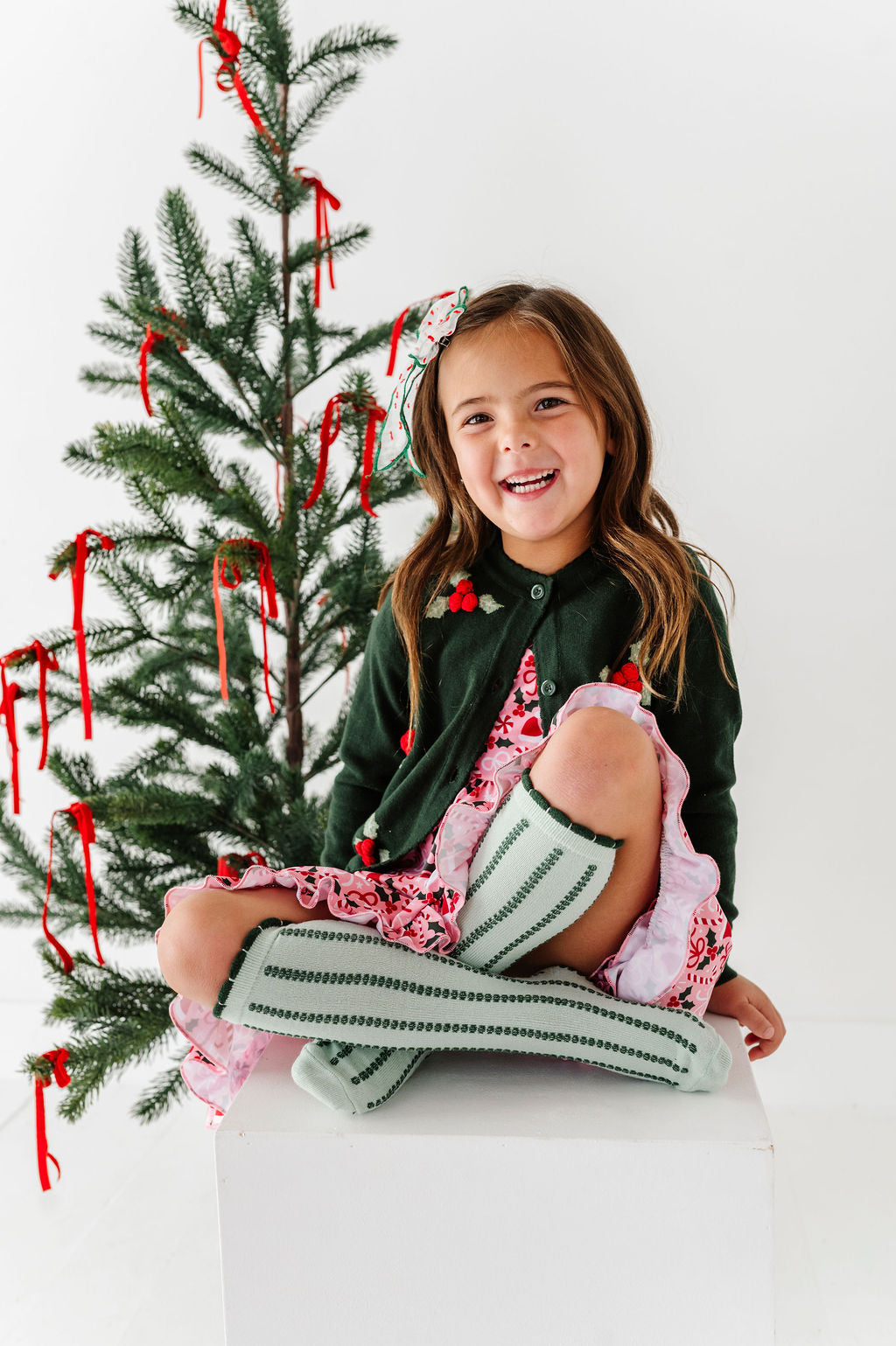 little girl sitting in front of christmas tree wearing christmas outfit with green vertical stripe knee high socks