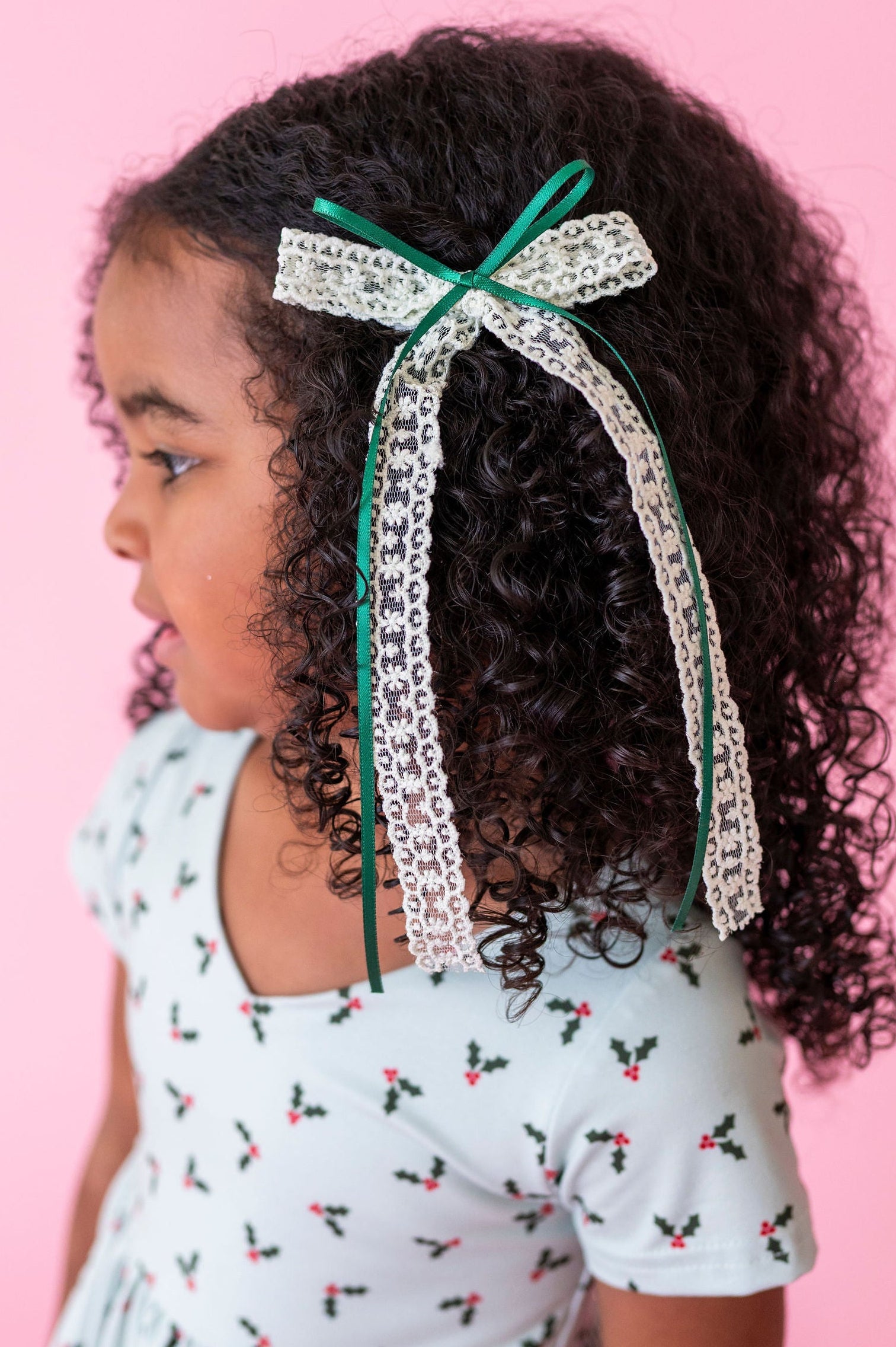 little girl in mint holly xmas dress with dark curly hair and mint and forest green lace and ribbon hair bow shown on a pink background