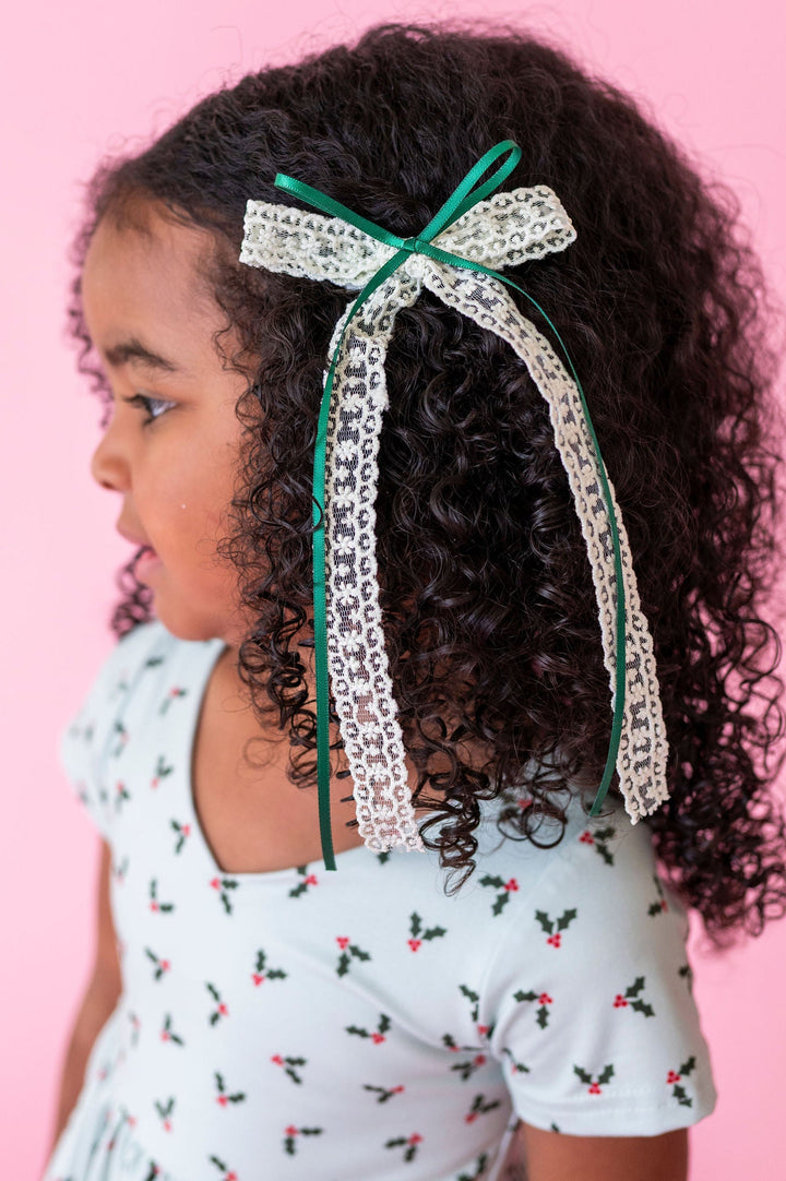 little girl in mint holly xmas dress with dark curly hair and mint and forest green lace and ribbon hair bow shown on a pink background