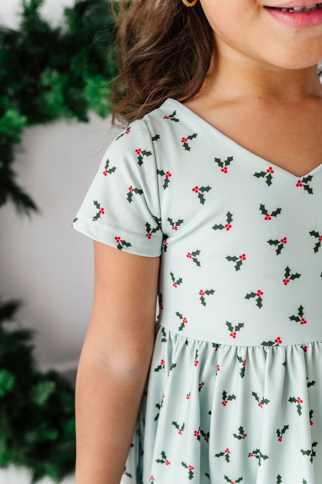 A close-up of a child wearing a short-sleeve V-neck dress with a light sage green base and a repeating holly and berries print in dark green and red. The background is a soft-focus holiday scene with green foliage.