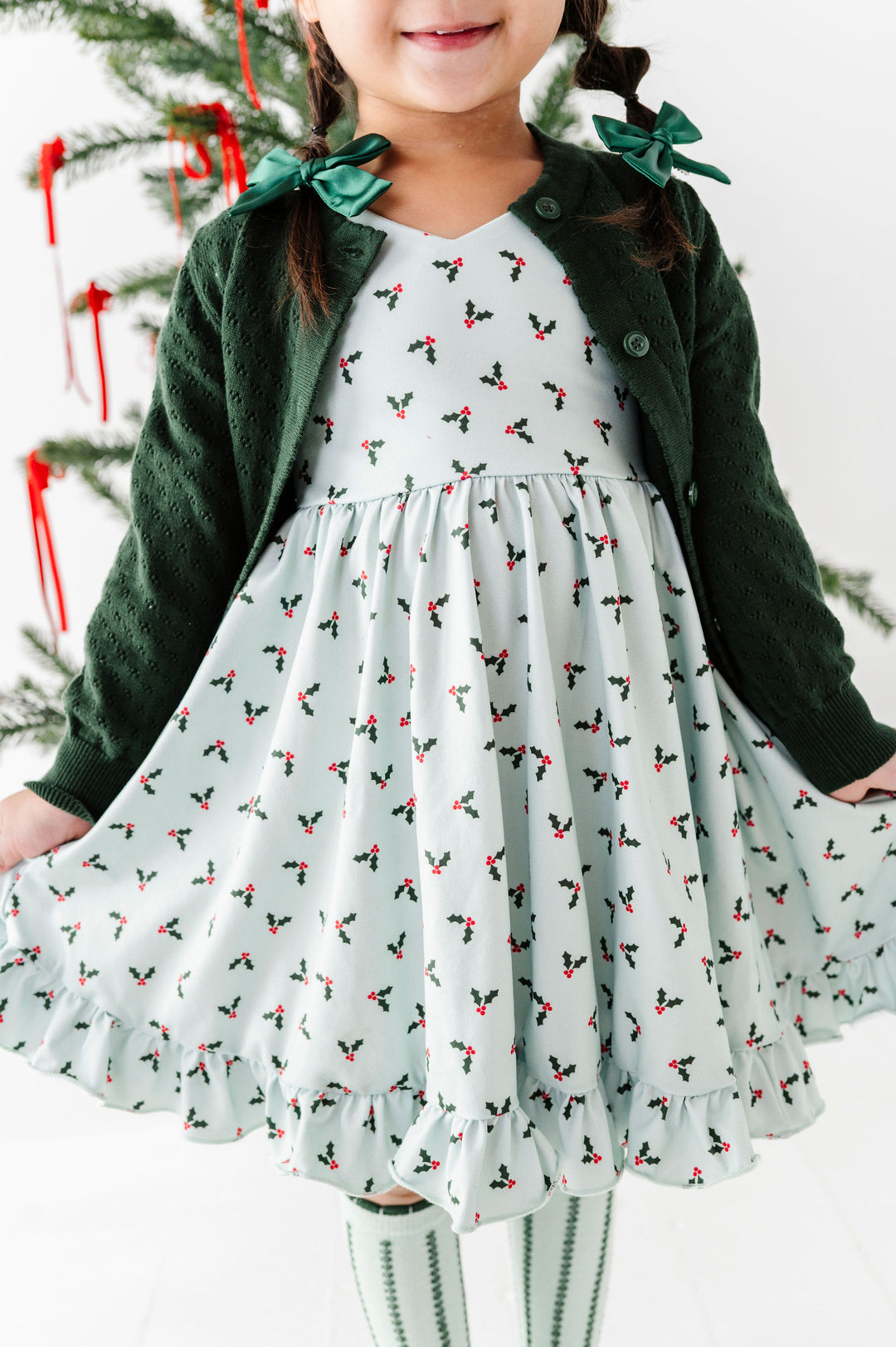 A young girl wearing a festive holiday dress with a dark green cardigan, featuring a white dress printed with small red and green holly motifs and green bow accents on the shoulders.