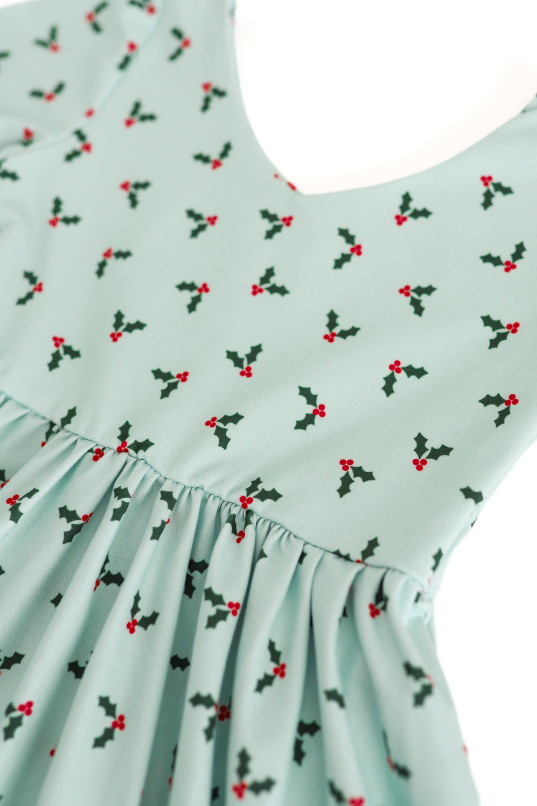 A close-up of a light mint green dress with a delicate all-over holly and berries print, featuring a gathered waist seam and v-neck design.