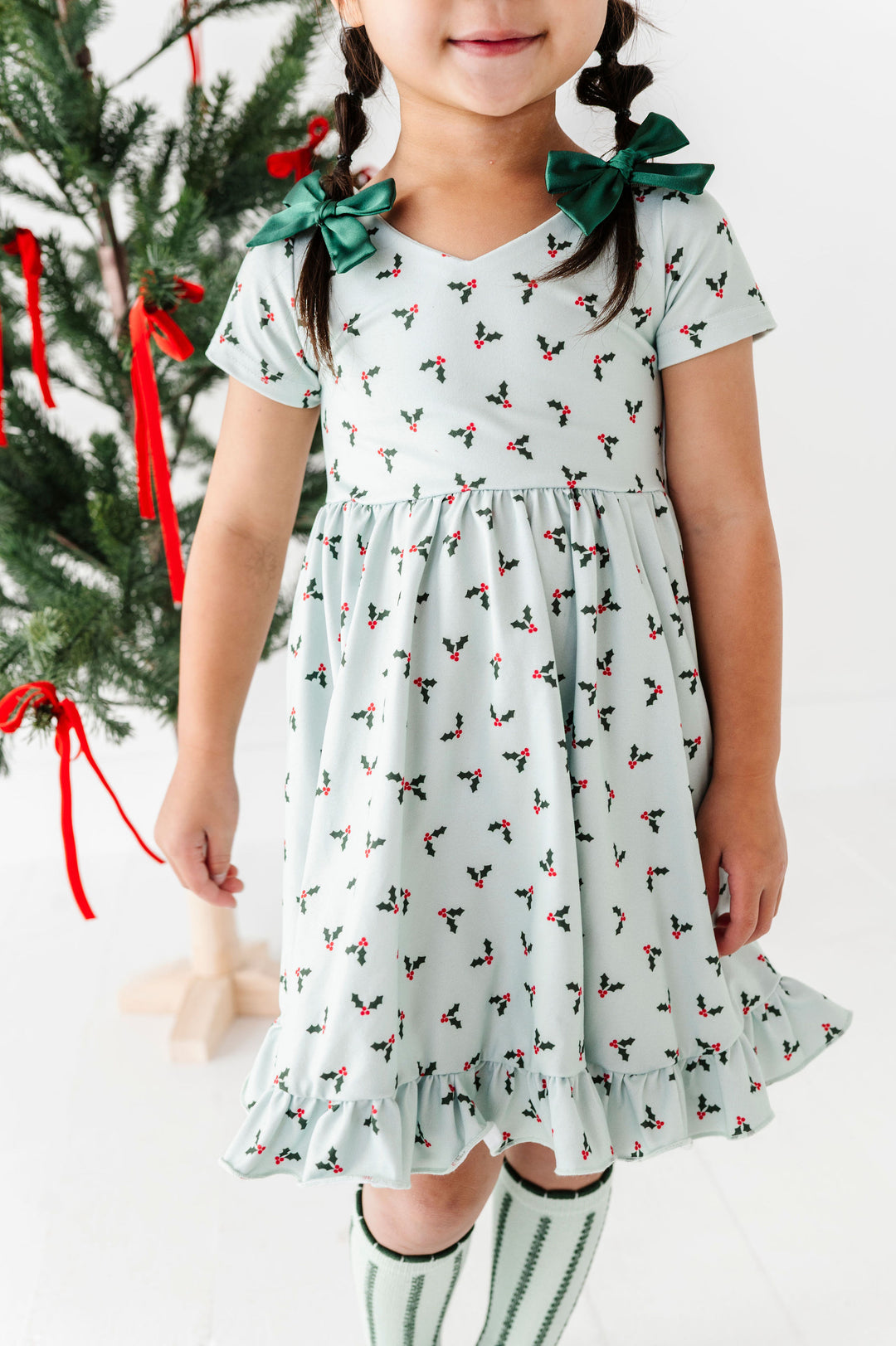 A young girl wearing a light sage green short-sleeve dress with a small red and green holly pattern, featuring dark green bow accents on the shoulders. She is standing in front of a decorated Christmas tree.