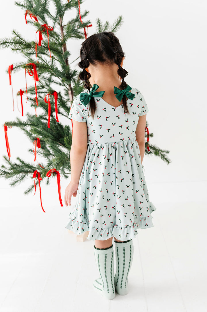 A young girl with braided hair is shown from behind wearing a light mint green dress with a small red and black pattern, green bow hair clips, and striped green socks. She is standing next to a decorated Christmas tree with red ribbons against a white background.