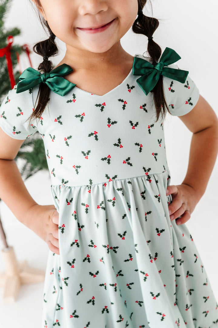 A young girl wearing a light mint green dress with a holly and berry print, featuring dark green bow accents on the shoulders. The photo is cropped from the chest up, showing her smiling and posing with hands on hips against a bright, clean indoor background.