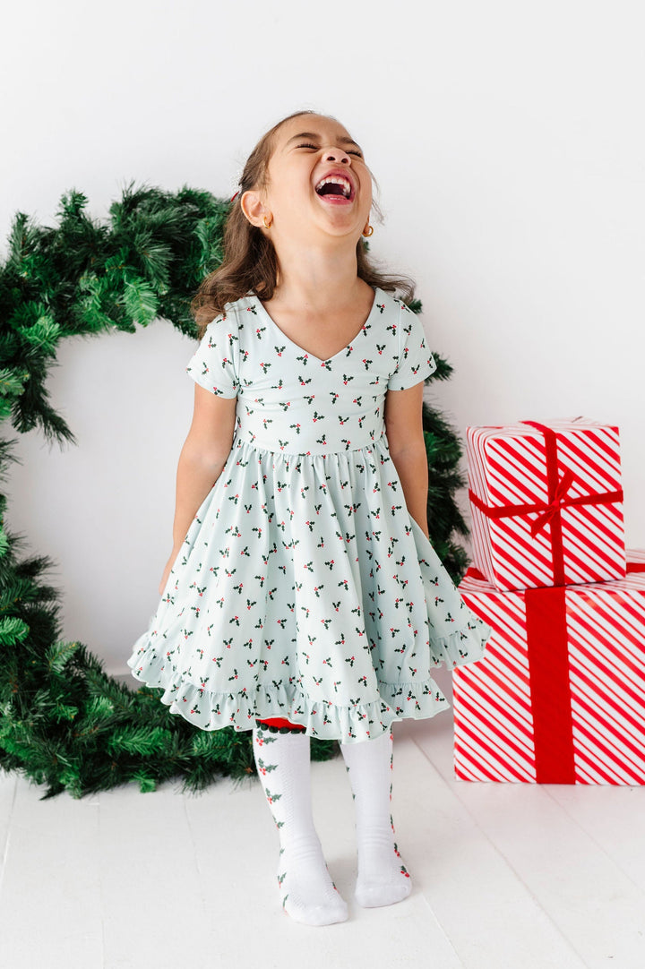 A young girl laughing joyfully in a mint green dress with a small green and red holly pattern, standing next to a green wreath and a stack of red and white wrapped presents against a white background.