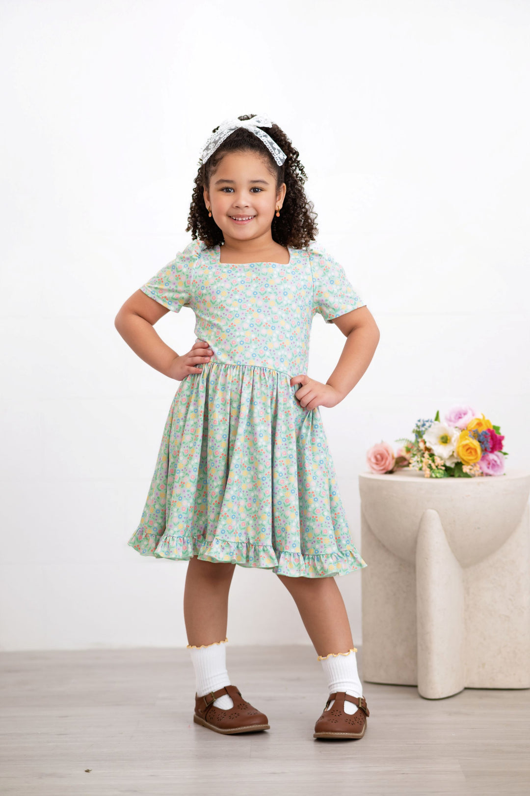 young girl in mint twirl dress with pastel floral print pared with white lace hair bow and white ribbed midi socks with yellow trim