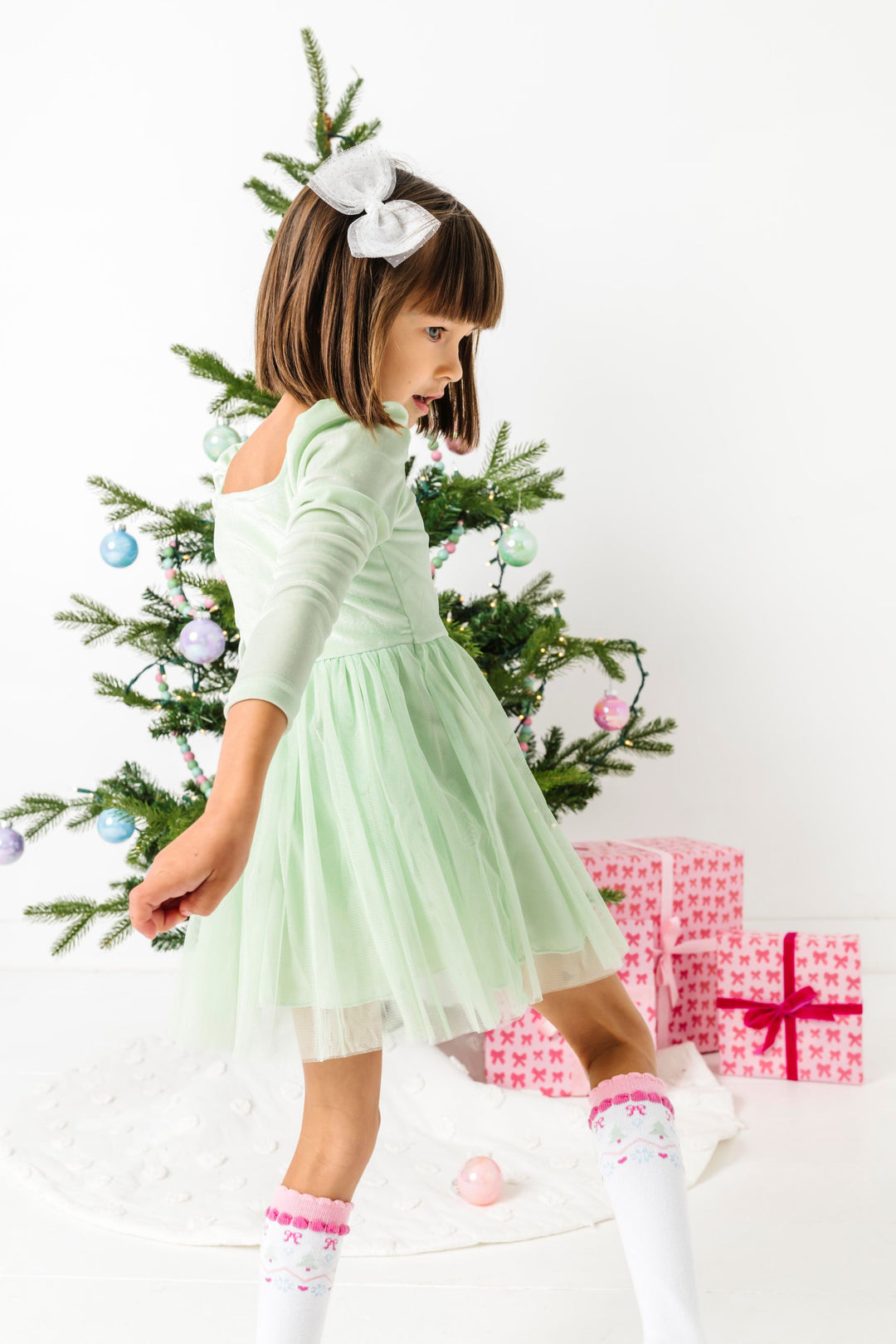 little girl in front of Christmas tree in mint green velvet long sleeve party dress with tulle skirt, pastel fair isle knee highs and sparkly white tutu nbow
