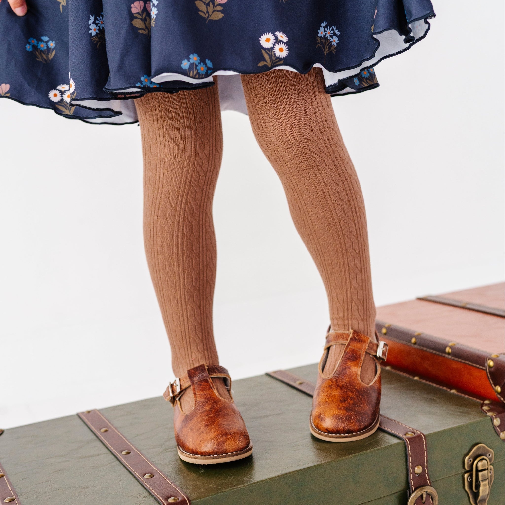little girl standing on vintage suitcase in light brown cable knit tights paired with navy blue floral twirl dress and brown mary janes