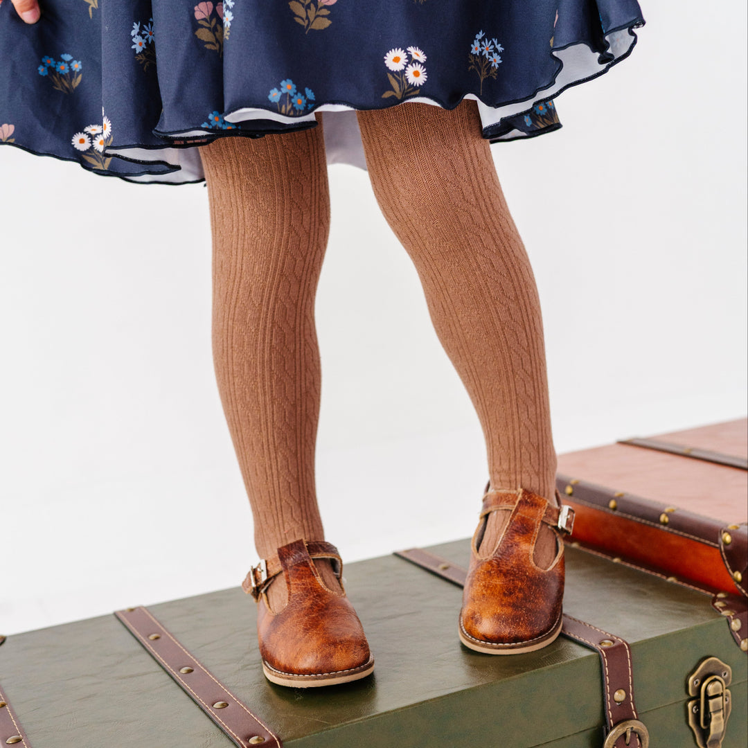 little girl standing on vintage suitcase in light brown cable knit tights paired with navy blue floral twirl dress and brown mary janes