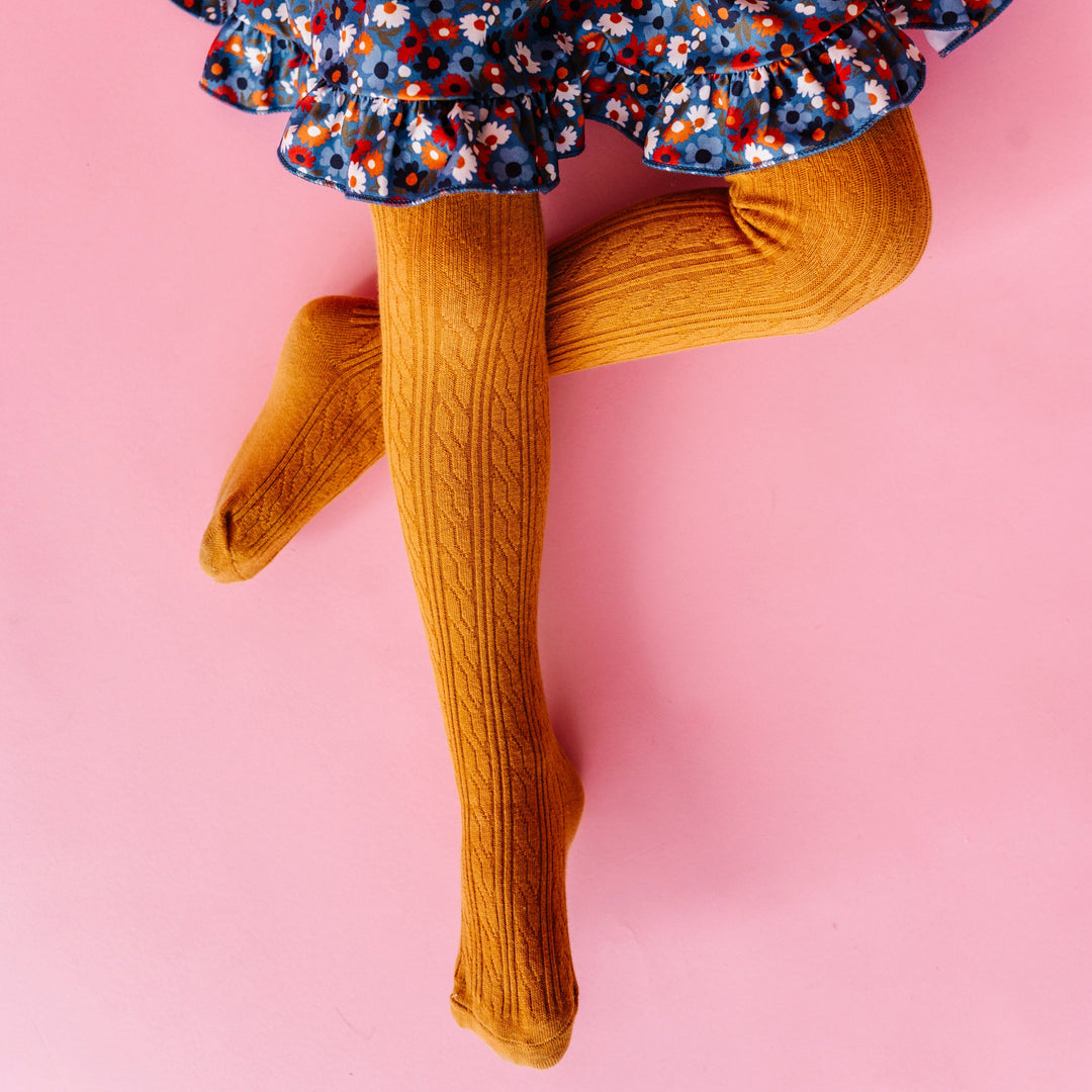 young girl sitting on pink backdrop in mustard cable knit tights paired with coordinating blue floral twirl dress