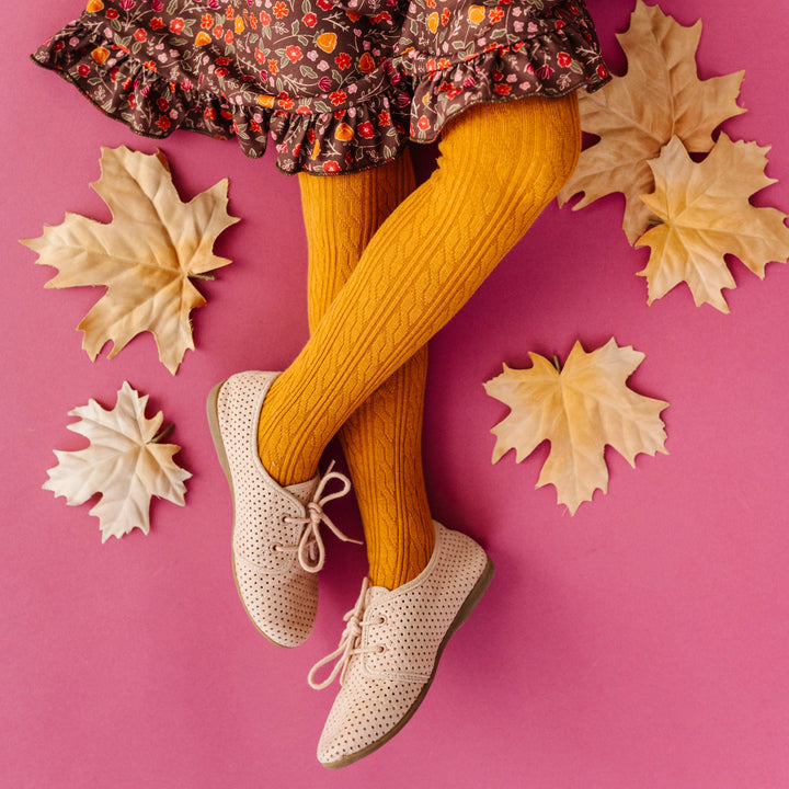 young girl sitting on pink backdrop with fall leaves wearing mustard cable knit tights paired with cream oxfords and brown floral twirl dress for fall
