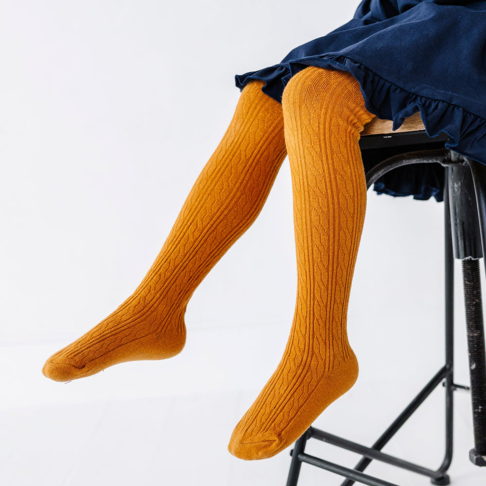 little girl sitting on chair wearing mustard cable knit tights paired with navy blue ruffle trim twirl dress