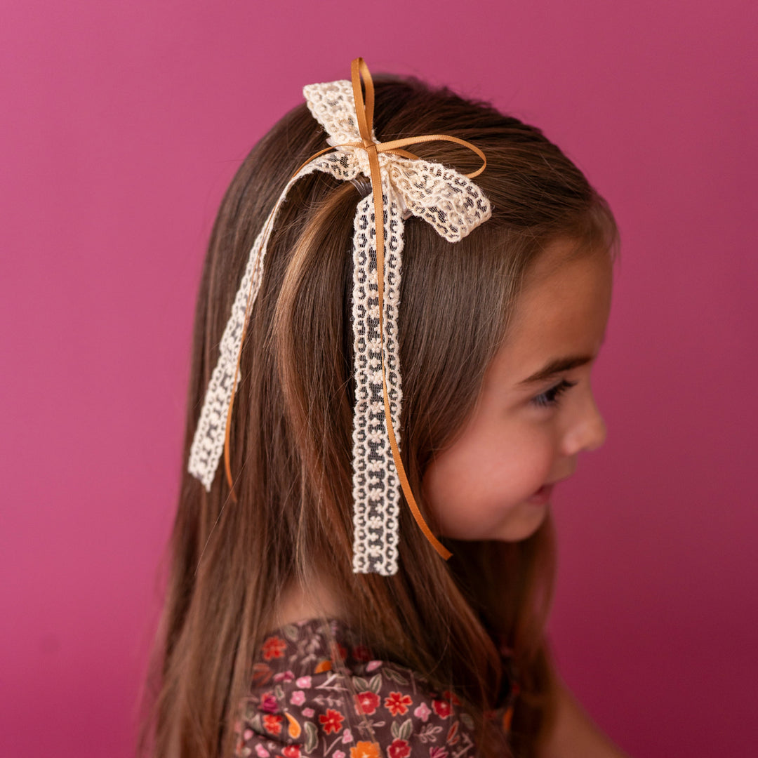 young girl in front of berry pink background wearing cream lace with mustard ribbon hair bow paired with brown fall floral dress